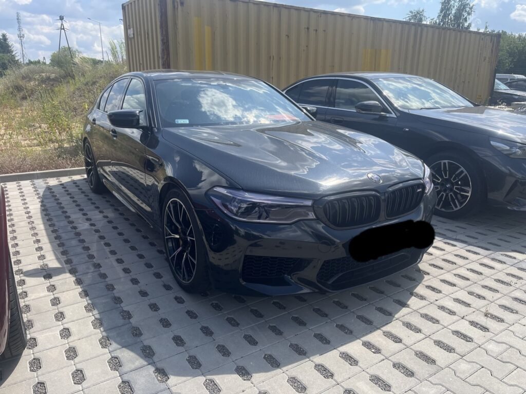 BMW M5 Competition 2018 Czarne widok od frontu w słoneczny dzień na samochód z zakrytą rejestracją.